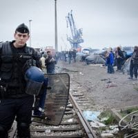 An armed police officer at Calais migrant camp. Photo by Squat le Monde (Photo Credit: Flickr)