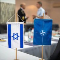 A NATO and an Israeli table side flags on a blurred buffet table and two military officers talking. Photo: NATO Maritime Command/File photo.