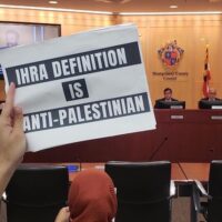 AN ACTIVIST HOLDS UP A SIGN AT A MONTGOMERY COUNTY COUNCIL MEETING ON OCTOBER 25 PROTESTING THE COUNCIL’S CONSIDERATION TO ADOPT THE IHRA DEFINITION OF ANTISEMITISM. (PHOTO: MARYLAND 2 PALESTINE/TWITTER)