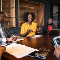 View between colleagues with focus on African professional in early 30s looking across conference table at young associate, smiling and gesturing as she speaks. (Photo by Johnny Greig)