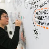 An East Asian man writes on a white board. To his right is a text bubble on the board that reads, in all capitals, "MACHINES WILL NEVER THINK AS HUMANS CAN."