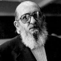 A black and white photograph of Paulo Freire later in life. Freire is bald, bearded, and wears large eyeglasses.