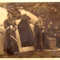Two Native women in traditional robes and beads stand outside the entrance to a hut-like structure on the Menominee Indian Reservation, crica 1913–18; nearby two other women are seated while another stands.