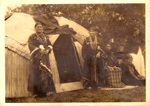 MR Online | Two Native women in traditional robes and beads stand outside the entrance to a hut like structure on the Menominee Indian Reservation crica 191318 nearby two other women are seated while another stands | MR Online