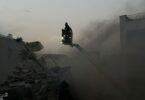 A person on a crane surveys the ruins of a bombed building, surrounded by smoke.