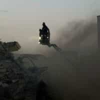 A person on a crane surveys the ruins of a bombed building, surrounded by smoke.