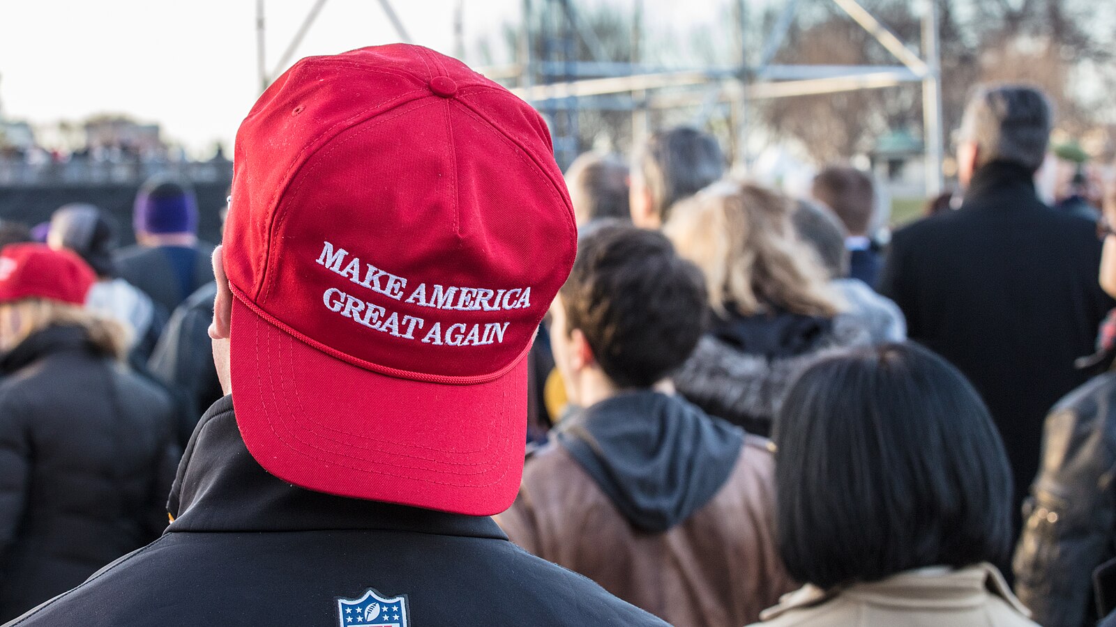 A crowd of people seen from behind; a person in the foreground wears a red Make America Great Again hat.