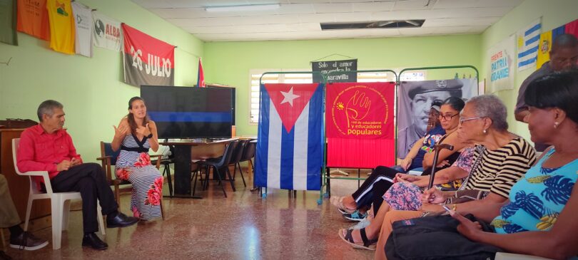 A group of people of various ages sit in a room decorated with Cuban Revolutionary memorabilia and posters. One person, wearing a long dress, is speaking while others listen.