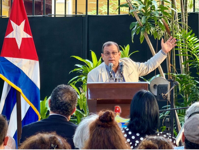 A middle-aged man stands at a podium, addressing an audience. A Cuban flag is visible to the left.
