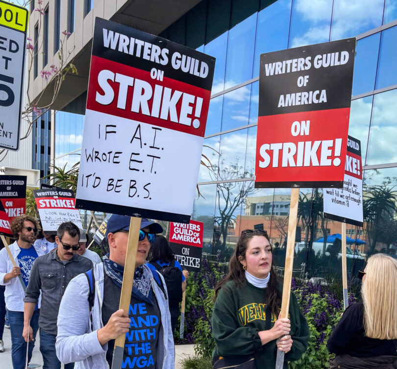 Striking WGA members walk a picket line in Los Angeles. In the foreground, a man holds a sign that reads, "If A.I. wrote E.T., it'd be B.S."