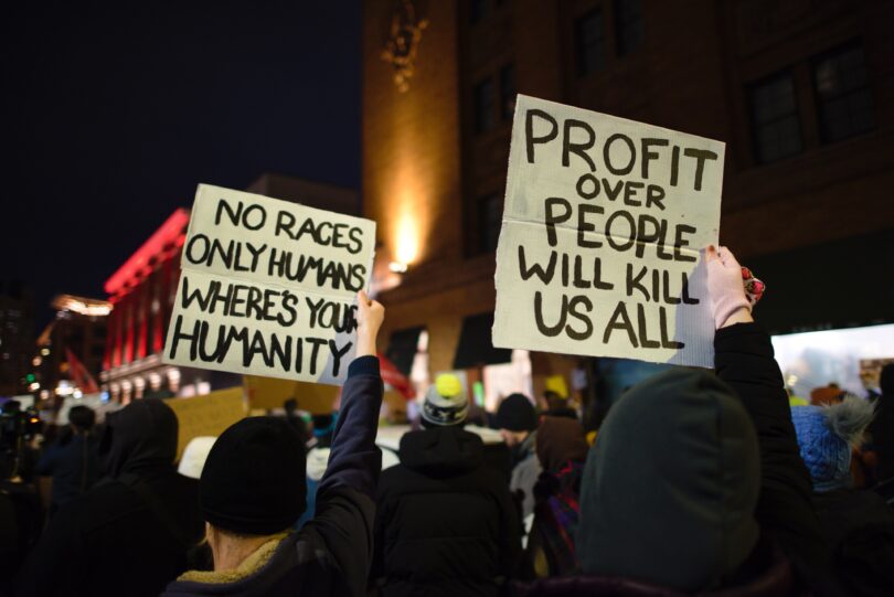 A crowd of protesters is seen from behind at night. Two people hold signs, one reading NO RACES ONLY HUMANS WHERE'S YOUR HUMANITY and another reading PROFIT OVER PEOPLE WILL KILL US ALL.
