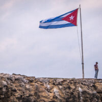 A man stands looking out at the edge of a rocky seaside precipice. Behind him, a large Cuban flag billows atop a flagpole.