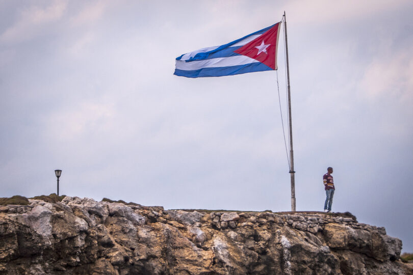 A man stands looking out at the edge of a rocky seaside precipice. Behind him, a large Cuban flag billows atop a flagpole.