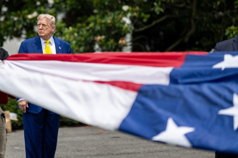 Part of a US flag in the foreground, with President Donald Trump standing in the background.