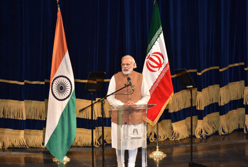 Indian Prime Minister Narendra Modi stands at a lectern onstage. Indian and Iranian flags are visible behind him.