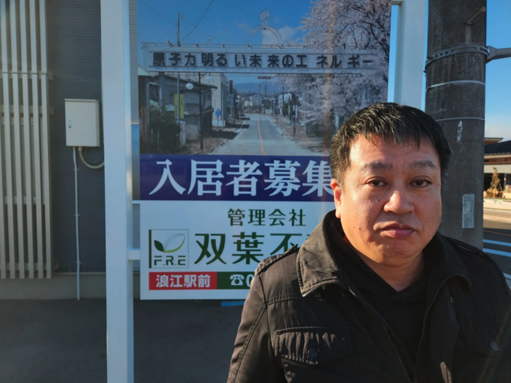 Yuji Onuma in front of his sign. The photo behind him shows the main street of Futaba, with the arch that Onuma designed as a schoolboy, which reads “Atomic power: Energy for a bright future.” Thomas A. Bass