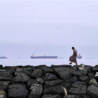 A man walks on rocks along the shore as oil tankers and cargo ships line up in the Strait of Hormuz, as seen from Khor Fakkan, United Arab Emirates, Wednesday, March 11, 2026. (AP Photo/Altaf Qadri)