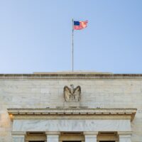 A frontal view of the top of the Federal Reserve headquarters. An eagle statue and US flag are visible.