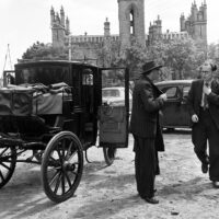 A black and white photo of two men in suits standing next to a carriage. A Gothic church is visible in the background.