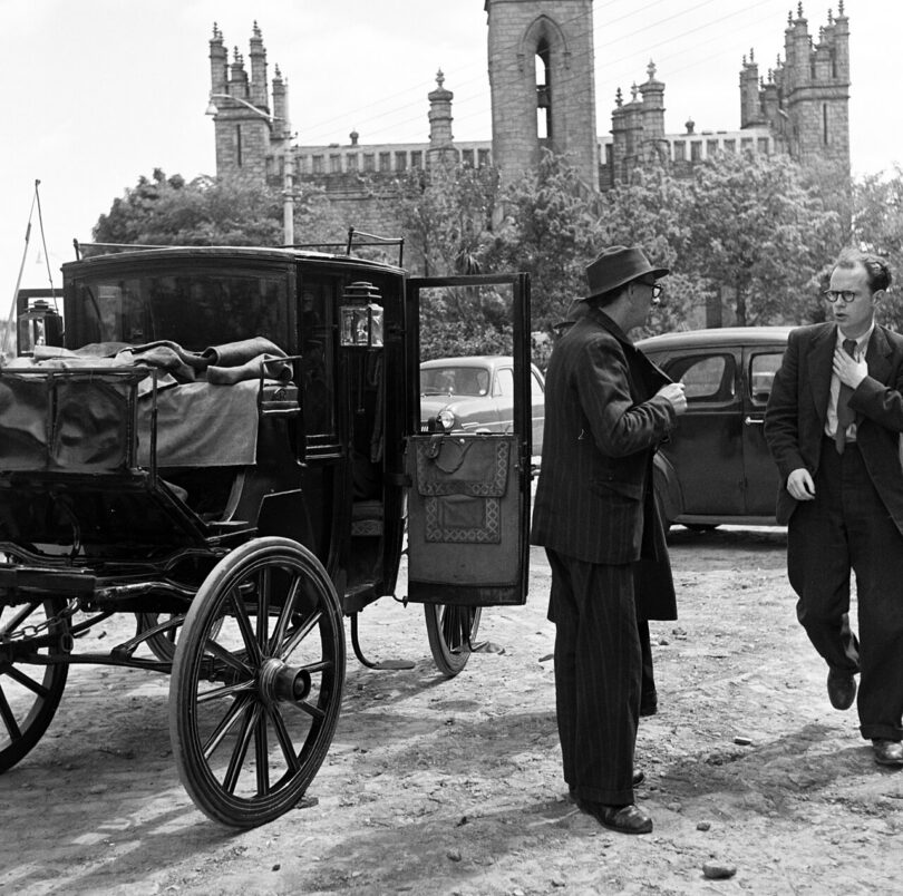 A black and white photo of two men in suits standing next to a carriage. A Gothic church is visible in the background.