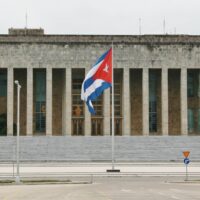 A wide short of the Palace of the Revolution in Havana, Cuba. A flagpole with a large Cuban flag stands at the foot of the building's front steps, in the center of the image.