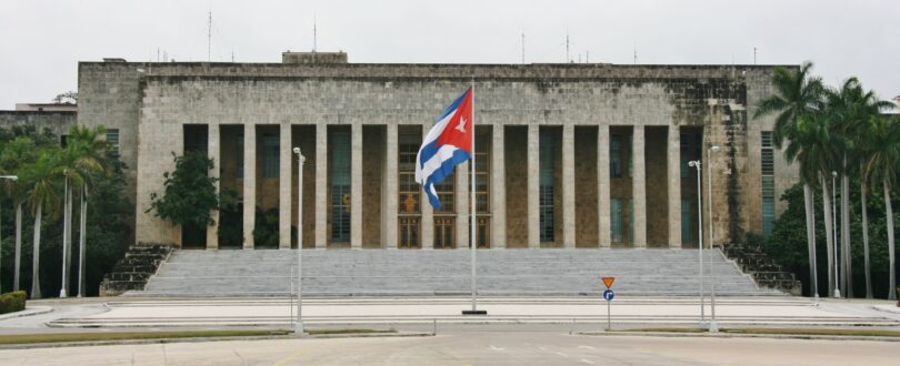 A wide short of the Palace of the Revolution in Havana, Cuba. A flagpole with a large Cuban flag stands at the foot of the building's front steps, in the center of the image.
