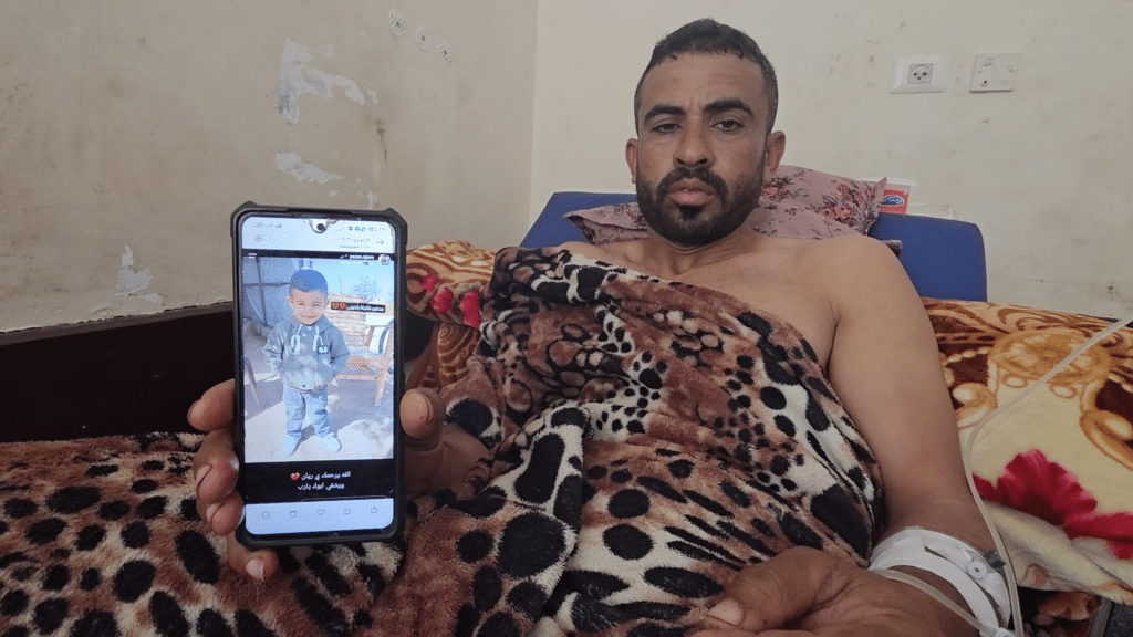 BAHAHA ABU AL-AJEEN AT AL-AQSA MARTYRS HOSPITAL WITH A PHOTO OF HIS SON, RAYYAN. (PHOTO: SCREENSHOT FROM VIDEO TESTIMONY GATHERED BY OSAMA KAHLOUT FOR MONDOWEISS)