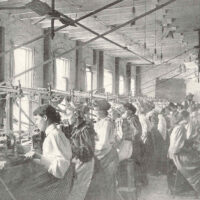 A row of women workers sit at machines used in the manufacture of buttons for clothing.
