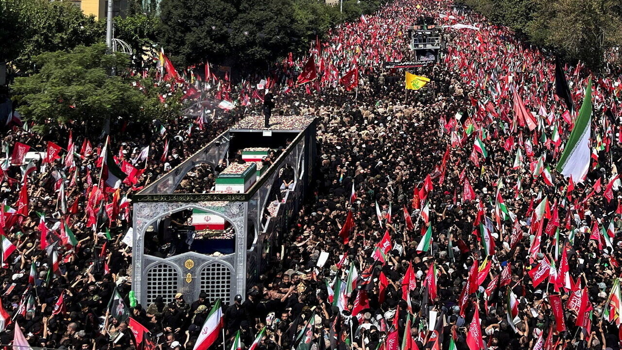 A vast crowd fills a street in Tehran, with many people waving Iranian flags.