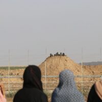 A row of women in hijabs are seen from behind, facing a group of Israeli soldiers on the other side of a border fence.