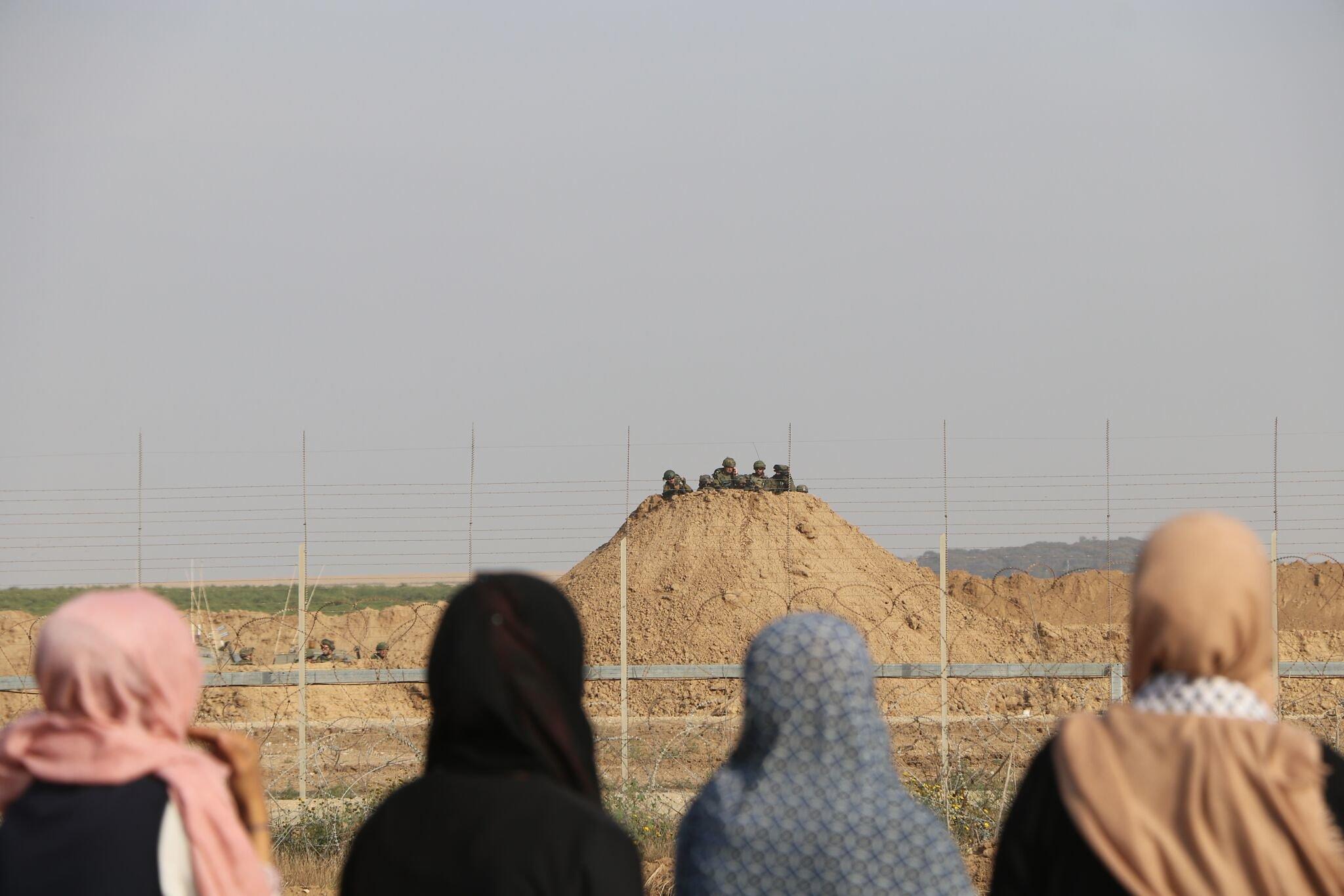 A row of women in hijabs are seen from behind, facing a group of Israeli soldiers on the other side of a border fence.