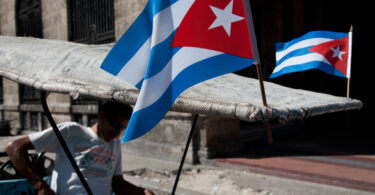 A man sits in a pedicab with two Cuban flags attached to its roof.