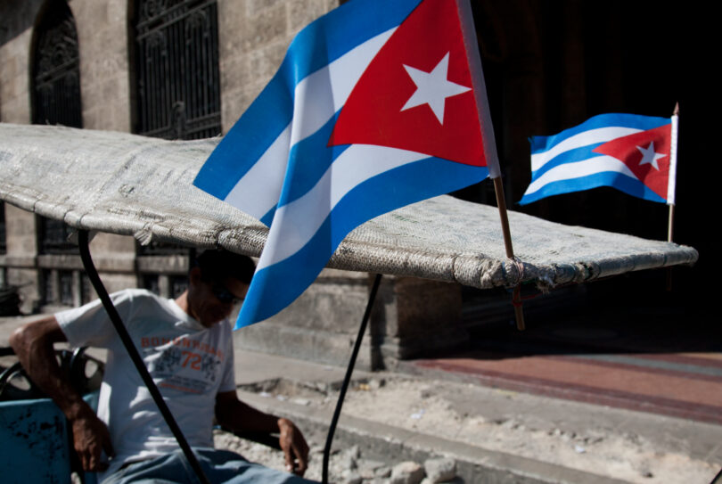 A man sits in a pedicab with two Cuban flags attached to its roof.