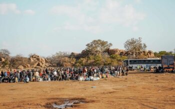 MIGRANT WORKERS GATHER WITH THEIR LUGGAGE BESIDE BUSES AT AN OUTDOOR DEPARTURE AND TRANSIT POINT, AWAITING ONWARD TRANSPORT IN LIMPOPO, SOUTH AFRICA (COPYRIGHT WECARE FOUNDATION, 2026)