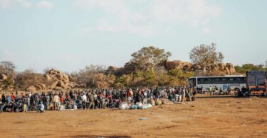 MIGRANT WORKERS GATHER WITH THEIR LUGGAGE BESIDE BUSES AT AN OUTDOOR DEPARTURE AND TRANSIT POINT, AWAITING ONWARD TRANSPORT IN LIMPOPO, SOUTH AFRICA (COPYRIGHT WECARE FOUNDATION, 2026)