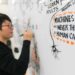 An East Asian man writes on a white board. To his right is a text bubble on the board that reads, in all capitals, "MACHINES WILL NEVER THINK AS HUMANS CAN."