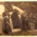 Two Native women in traditional robes and beads stand outside the entrance to a hut-like structure on the Menominee Indian Reservation, crica 1913–18; nearby two other women are seated while another stands.